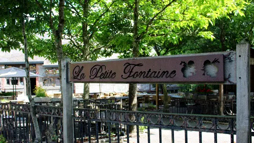 Enseigne en bois et métal du restaurant La Petite Fontaine à Achouffe, entourée d'arbres et d'une terrasse extérieure