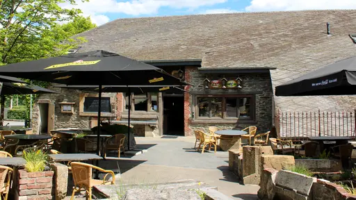 Grande terrasse ensoleillée du restaurant La Petite Fontaine à Achouffe avec tables rondes, fauteuils en rotin et grands parasols noirs devant une bâtisse en pierre d'Ardenne